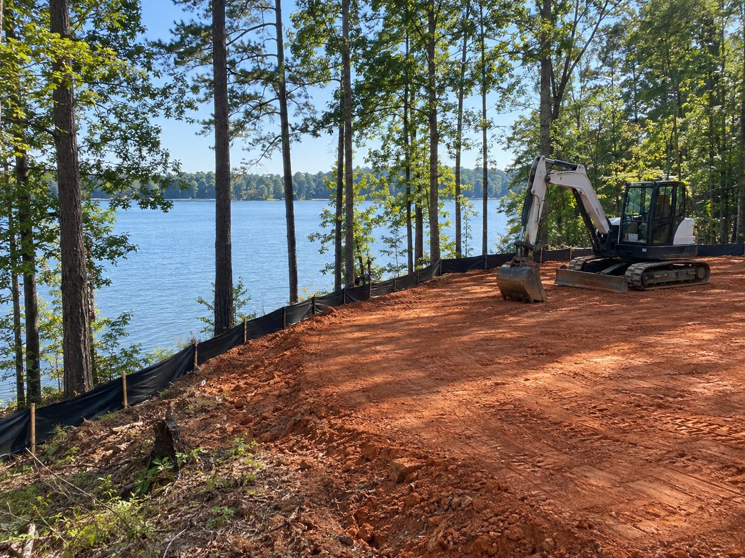 Excavator grading a red clay building pad on a wooded lake lot with silt fence installed